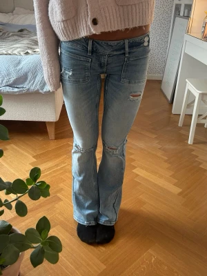 Gina 14+ jeans - Snygga ljusblå bootcut jeans med slitningar på knäna och låret. Jeansen har framfickor med lock och markerade sömmar. Perfekta för en avslappnad och trendig look. Materialet är klassisk denim i bomull.