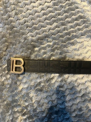 Svart Burberry bälte med B-spänne - Snyggt svart bälte från Burberry med klassiskt rutmönster och stort B-format metallspänne. Bältet är gjort i skinn och har det ikoniska Burberry-märket präglat på remmen. Perfekt accessoar för att lyfta din outfit.