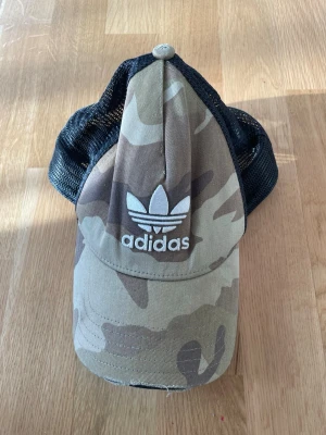 Camo keps från Adidas Originals - Säljer en snygg keps från Adidas Originals med klassiskt kamouflagemönster i beige, brunt och grönt. Framsidan har broderad vit Adidas-logga och baksidan är i svart mesh för extra ventilation. Justerbar passform och böjd skärm.