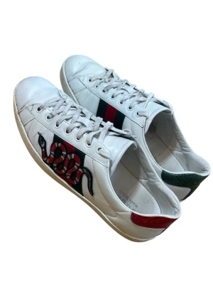 Gucci vita sneakers med orm och ränder - Säljer ett par vita Gucci sneakers med ikoniska gröna och röda ränder på sidan samt en broderad orm. Skorna har snörning, rund tå och detaljer i skinn. Baktill finns en grön och en röd hälflik för extra stilpoäng.