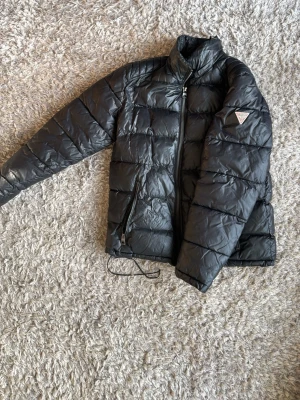 Svart dunjacka från Guess - Snygg svart dunjacka från Guess med hög krage och dragkedja framtill. Jackan har två fickor med dragkedja och den klassiska Guess-loggan på vänster ärm. Perfekt för kalla vinterdagar och har en stilren, quiltad design.
