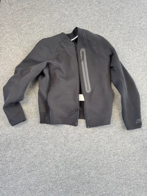 Svart träningströja från Nike - Snygg svart tröja från Nike med dragkedja framtill och en vertikal ficka med dragkedja på bröstet. Jackan har en minimalistisk design och är tillverkad i ett mjukt syntetmaterial. Perfekt för dig som gillar sportig och stilren look.