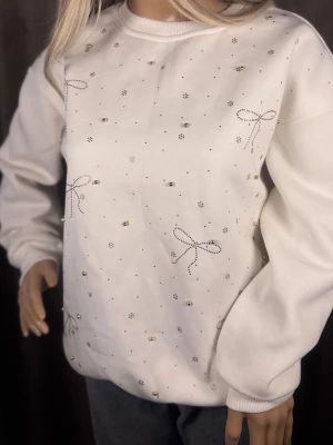 Vit sweatshirt med pärlor och rosetter - Superfin vit sweatshirt med små guldfärgade pärlor och söta rosett-mönster i strass. Tröjan har rund halsringning och ribbade muddar vid ärmslut och nederkant. Perfekt för dig som gillar detaljer och en söt, stilren look.