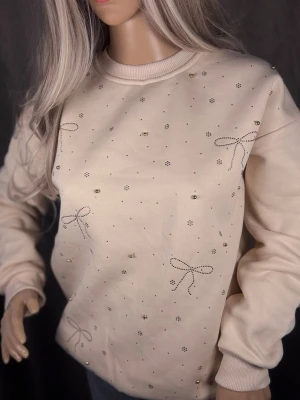 Beige sweatshirt med pärlor och rosetter - Supersöt beige sweatshirt med små pärlor och dekorativa rosetter i silverfärgade nitar. Tröjan har rund halsringning, ribbade muddar och är gjord i mjukt bomullsmaterial. Perfekt för dig som gillar detaljer och en feminin touch på din outfit.