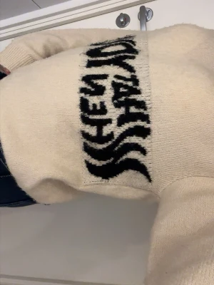 Beige stickad tröja med text - Stickad tröja i beige med svart text över bröstet. Tröjan har rund halsringning och långa ärmar. Mjuk och skön modell som passar perfekt till jeans. 