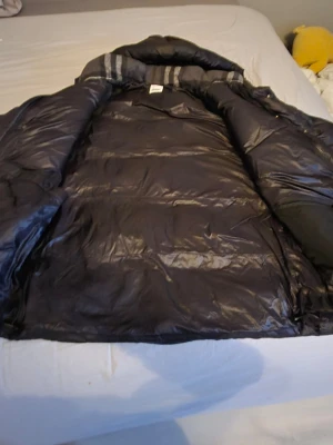 Svart dunjacka från Burberry - Snygg svart dunjacka från Burberry med klassisk design och hög krage. Jackan har en glansig finish och är perfekt för kalla vinterdagar. Märket Burberry syns tydligt på insidan. Passar dig som vill ha både stil och värme. Pris kan diskuteras 