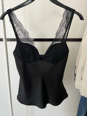 Svart linnetopp med spets från Zara - Andvänd endast en gång. Supersnygg svart linnetopp från Zara med spetsdetaljer på axelbanden och över bysten. Sidenliknande material ger en lyxig känsla och toppen har en figurnära passform. Perfekt att styla med jeans eller kjol för en trendig look. 