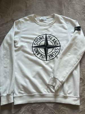 Vit sweatshirt från Stone Island - Säljer en vit sweatshirt från Stone Island med stort broderat logotyptryck fram på tröjan och klassisk patch på ärmen. Tröjan har rund halsringning och är gjord i mjuk bomull. Perfekt för dig som gillar en fancy look och snygga detaljer. Som man kan se på sista bilden så börjar det att spricka upp lite men det är enkelt att fixa! Pris kan diskuteras vid snabb affär!