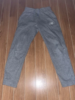 Gråa mjukisbyxor från Nike - Säljer ett par gråa mjukisbyxor från Nike med klassisk logga på vänster ben. Byxorna har resår i midjan och muddar vid bensluten för en skön passform. Perfekta för chill dagar eller träning. Materialet är mjukt och bekvämt, troligen bomullsblandning. Storlek 147-158. Bara hör av er vid funderingar!😃