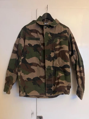 Camouflage vindjacka med dragkedja - Säljer en grön, brun och beige camouflage vindjacka med dragkedja framtill och två bröstfickor. Jackan har klassisk krage, långa ärmar och justerbar nederkant. Perfekt för dig som gillar streetwear och utility-vibe. 