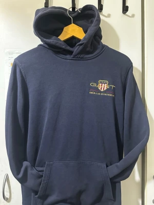 Mörkblå hoodie från GANT - Mörkblå hoodie från GANT med broderad logga och texten 'American Sportswear' på bröstet. Klassisk känguruficka framtill och justerbar huva. Tillverkad i mjukt bomullsmaterial som är skönt att bära. Perfekt för en avslappnad och sportig stil.