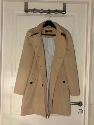 Beige trenchcoat med svarta knappar - Stilren beige trenchcoat med klassisk dubbelknäppning och mörkbruna knappar. Kappan har två fickor framtill och axelklaffar för en tidlös look. Insidan är fodrad med ljusrandigt tyg. Perfekt för dig som vill ha en snygg och enkel trench till vardagslooken.