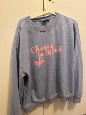 Ljuslila sweatshirt Cherry Bomb - Ljuslila sweatshirt med trycket 'Cherry Bomb' i rosa och motiv av körsbär med rosett på bröstet. Tröjan har rund hals, ribbade muddar och avslappnad passform. Perfekt för en chill och trendig look.