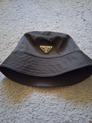 Svart bucket hat från Prada - Snygg svart bucket hat från Prada i återvunnen nylon med klassisk logga framtill. Perfekt accessoar för att lyfta din stil och ge en trendig touch till outfiten. Hatten har en mjuk och följsam form som sitter bekvämt på huvudet.