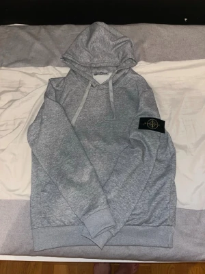Grå hoodie från Stone Island - Snygg grå hoodie från Stone Island med klassisk patch på vänster ärm. Hoodien har dragsko i huvan och ribbade muddar vid ärmslut och nederkant. Tillverkad i mjukt bomullsmaterial som är perfekt för chill dagar.