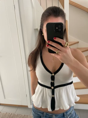 Vit volangtopp med svarta detaljer - Superfin vit ribbad topp med svarta kanter och guldknappar framtill. Toppen har v-ringning, markerad midja och volangig nederdel. Från Zara, går inte längre att köpa. 