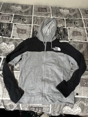 Grå och svart hoodie från The North Face - Snygg hoodie med dragkedja från The North Face i grått och svart. Stor huva med dragsko, två fickor fram och logga på bröstet. Tillverkad i mjukt material, perfekt för chill eller streetwear. Klassisk färgkombination och stilren design.
