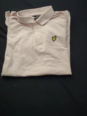Ljusrosa pikétröja från Lyle & Scott - Säljer en ljusrosa pikétröja från Lyle & Scott med klassisk krage och tre knappar framtill. Tröjan har det ikoniska gula logomärket broderat på bröstet och är tillverkad i mjuk bomull. Perfekt för en clean och stilren look.