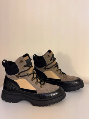 Beige och svarta kängor från Agnes Cecilia - Snygga kängor från Agnes Cecilia i beige mocka och svart läder med grov sula. De har detaljer i guld, svarta snören och mesh vid ankeln för extra komfort. Perfekta för dig som vill ha en cool och robust look till hösten eller vintern. På kängan står storlek 40 men det är 39, 25cm invändigt. Inte för breda fötter. 