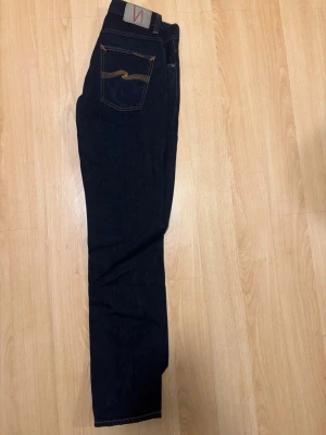 Mörkblå jeans från Nudie Jeans - Säljer ett par mörkblå jeans från Nudie Jeans med klassisk femficksdesign och kontrastsömmar. Jeansen har rak passform och är tillverkade i kraftig denim. Snygg detalj med Nudie-loggan på bakfickan och patch i midjan. Modellen är Grim Tim och storlek 31/34. 