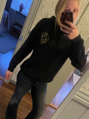 Svart hoodie med coolt tryck - Svart hoodie med huva och snörning, dekorerad med ett gult smiley-tryck på bröstet och ett stort gult motiv med text på ryggen. Perfekt för en avslappnad och cool stil.