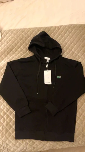 Svart hoodie från Lacoste med dragkedja - Svart hoodie från Lacoste med dragkedja framtill, huva med dragsko och den klassiska gröna krokodilloggan broderad på bröstet. Regular fit och ribbade muddar vid ärmslut och nederkant. Perfekt för en chill och stilren look.
