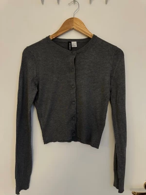 Mörkgrå cropped kofta från H&M - Snygg mörkgrå cropped kofta från H&M med rund halsringning och knappar framtill. Tillverkad i mjukt, tunt stickat material och har långa ärmar samt ribbade muddar vid ärmslut och nederkant. Perfekt att styla över ett linne eller t-shirt.