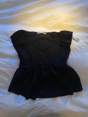 Svart volangblus med v-ringning - Snygg svart blus med v-ringad hals och volangdetaljer vid axlarna. Toppen har en lös passform nedtill och korta ärmar. Använd 1-2 gånger.