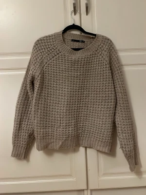 Beige stickad tröja från Gina Tricot - Mysig beige stickad tröja från Gina Tricot med grovstickat mönster och rund halsringning. Tröjan har långa ärmar och ribbade muddar vid ärmslut och nederkant. Perfekt för lager-på-lager och chill dagar.