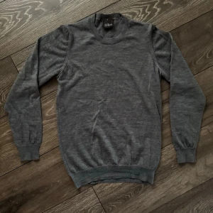 Oscar Jacobson tröja - En stilren grå stickad tröja från oscar Jacobson med rund halsringning och långa ärmar. Tröjan har ribbade muddar vid ärmslut och nederkant. Perfekt för lager-på-lager och enkel att matcha med andra plagg.