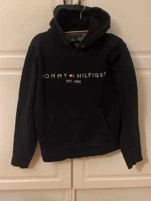 Svart hoodie Tommy Hilfiger - Svart hoodie från Tommy Hilfiger med broderad logga och text 'EST. 1985' på bröstet. Klassisk känguruficka framtill och justerbar huva med metallöljetter. Mjuk bomullsblandning och ribbade muddar för extra komfort.