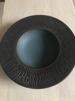 Djup tallriksmodell uppläggningsfat från Danmark i keramik - Säljer en djup tallrik  mer som en dekoration till ex frukt i keramik med matt mörkblå insida och svart, räfflad kant. Tallriken har en stilren och modern design, perfekt för soppor eller pastarätter. Märkt undertill med 'Danmark'.