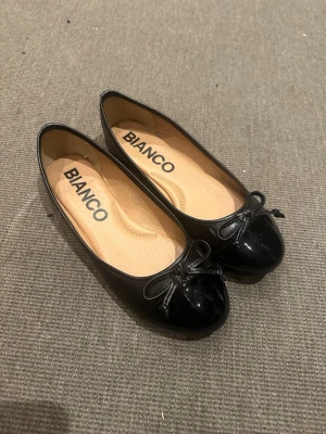 Svarta ballerinaskor från Bianco strl 40 - Klassiska svarta ballerinaskor från Bianco i storlek 40. Skorna har rund tå, liten rosett framtill och är tillverkade i syntetmaterial med en blank finish. Perfekta till jeans eller kjol för en enkel och stilren look.
