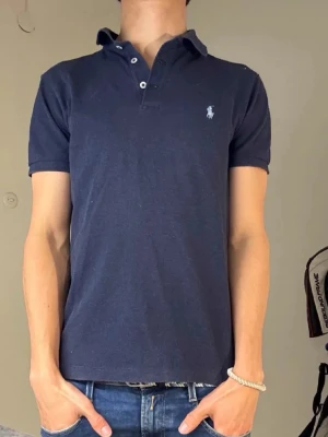 Mörkblå piké från Ralph Lauren - Klassisk mörkblå pikétröja från Ralph Lauren med korta ärmar och vit broderad logga på bröstet. Tröjan har krage och knappar framtill, tillverkad i mjuk bomull som känns skön mot huden. Perfekt till jeans eller shorts för en clean look.