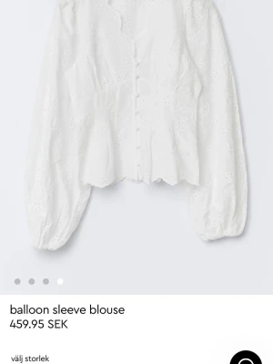 Vit blus med ballongärmar och spets - Söt vit blus med ballongärmar och dekorativa knappar framtill. Blusen har broderade spetsdetaljer och vågig nederkant som ger en romantisk känsla. Perfekt att styla med jeans eller kjol för en trendig look.