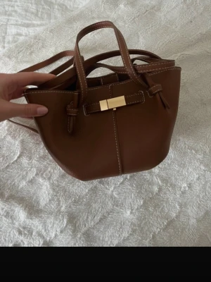 Brun handväska  - Snygg brun minihandväska i slätt läder med vita sömmar och guldfärgat spänne framtill. Väskan har både handtag och justerbar axelrem, perfekt för att bära crossbody. Stilren och trendig modell med detaljer i minimalistisk design.