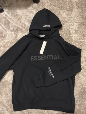 Svart Essentials hoodie - Svart hoodie från Essentials med stor logga tryckt på bröstet och mindre tryck på ärmen. Har kvar lappen.