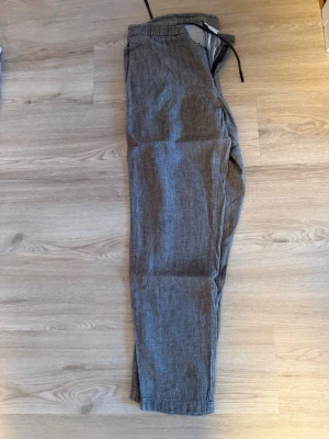Mörkgröna/gråa  linnebyxor med dragsko - Snygga grå linnebyxor med avslappnad passform och elastisk midja med dragsko. Byxorna har sidfickor och är tillverkade i ett lätt och luftigt linnetyg, perfekta för en chill och stilren look. Passar perfekt för dig som är runt 180 cm!