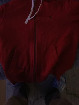 Röd hoodie med dragkedja och fickor - Röd hoodie med hel dragkedja framtill, vita snören i huvan och två stora fickor. Klassisk och enkel design, perfekt för en avslappnad stil. Liten broderad logga på bröstet.