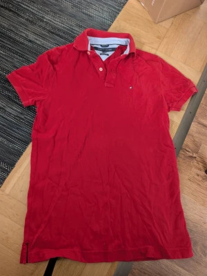 Röd pikétröja från Tommy Hilfiger stl M - Snygg röd pikétröja från Tommy Hilfiger i modellen Custom Fit. Tröjan har klassisk krage, knappar vid halsen och en liten logga på bröstet. Tillverkad i bomull för en bekväm känsla och passar perfekt till en avslappnad men stilren look.