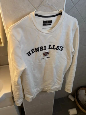 Vit sweatshirt från Henri Lloyd - Vit sweatshirt från Henri Lloyd med svart broderad logga och brittisk flagga på bröstet. Rund halsringning och klassisk passform. Perfekt för en clean och stilren look.