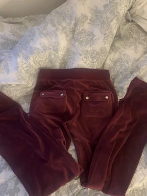 Vinröda velourbyxor Juicy Couture XS - Mysiga vinröda mjukisbyxor från Juicy Couture i velour. Byxorna har två bakfickor med knappar och bred resår i midjan. Perfekta för en chill och trendig look.