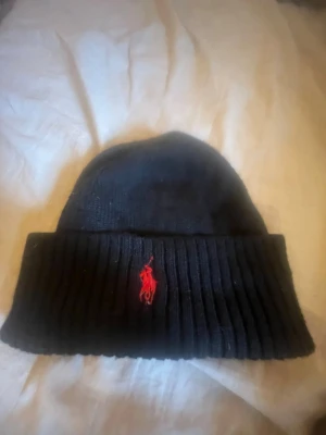 Svart ribbstickad mössa från Polo Ralph Lauren - Svart ribbstickad mössa från Polo Ralph Lauren med uppvikt kant och klassiska röda logotypen broderad framtill. Mjuk och stretchig, perfekt för kalla dagar. Enkel och stilren design som funkar till det mesta.