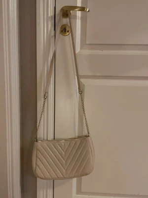Beige quiltad axelväska med kedja - Stilren beige axelväska med quiltat mönster och guldfärgad kedjedetalj på axelremmen. Väskan har dragkedja upptill och ett innerfack med dragkedja. Perfekt storlek för det viktigaste och lätt att matcha till olika outfits.