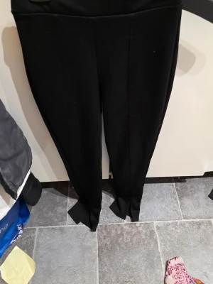 Svarta leggings med slits nertill - Snygga svarta leggings med hög midja och slitsar nertill på benen. De har en söm framtill som ger en dressad känsla. Perfekta att matcha med oversized tröjor eller crop tops. Materialet är stretchigt och följsamt för en bekväm passform.