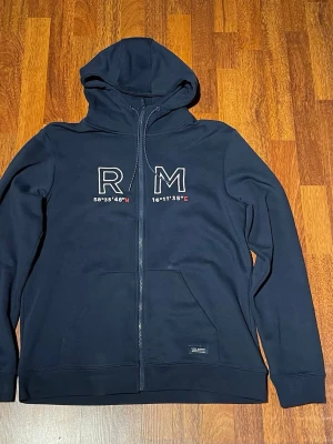 Marinblå hoodie med dragkedja RM - Snygg marinblå hoodie från Race Marine med dragkedja framtill och stor huva. På bröstet står det 'RM' och koordinater broderat i vitt och rött. Praktisk känguruficka och diskret Sail Racing-märke nedtill. Perfekt för en avslappnad streetwear-look. Max andvänd 3 gånger. Pris kan diskuteras!