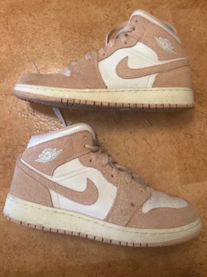 Beige och vita Air Jordan 1 Mid - Säljer ett par Air Jordan 1 Mid sneakers i beige mocka och vitt läder. Klassisk Nike Swoosh på sidan och Air Jordan-logga vid ankeln. Skorna har en vit mellansula och beige yttersula. Perfekta för dig som gillar streetwear och snygga detaljer. Använda 1 gång!