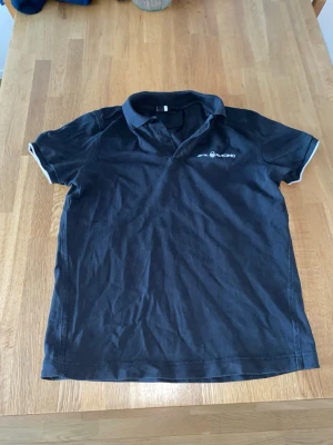 Svart pikétröja från Sail racing - Klassisk svart pikétröja från Sail racing med vit logga på bröstet och tryck på ryggen. Tröjan har korta ärmar med vita detaljer vid ärmsluten och krage. Perfekt till en avslappnad stil och enkel att matcha med jeans eller shorts.