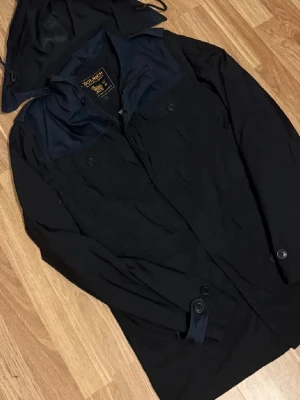 Svart regnjacka från Woolrich - Snygg svart regnjacka från Woolrich med avtagbar huva och knäppning framtill. Jackan har klassisk design med knappar vid ärmsluten och hög krage. Perfekt för regniga dagar och enkel att matcha med olika outfits.