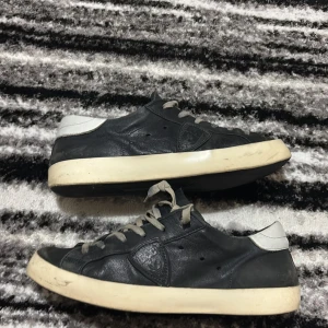 Phillipe model  - Snygga svarta sneakers från Philippe Model med vit häl och grå snörning. Skorna har en rund tå, platt sula och är tillverkade i skinn med märkets logga på sidan. Perfekta för dig som gillar stilrena och klassiska sneakers med exklusiv känsla.